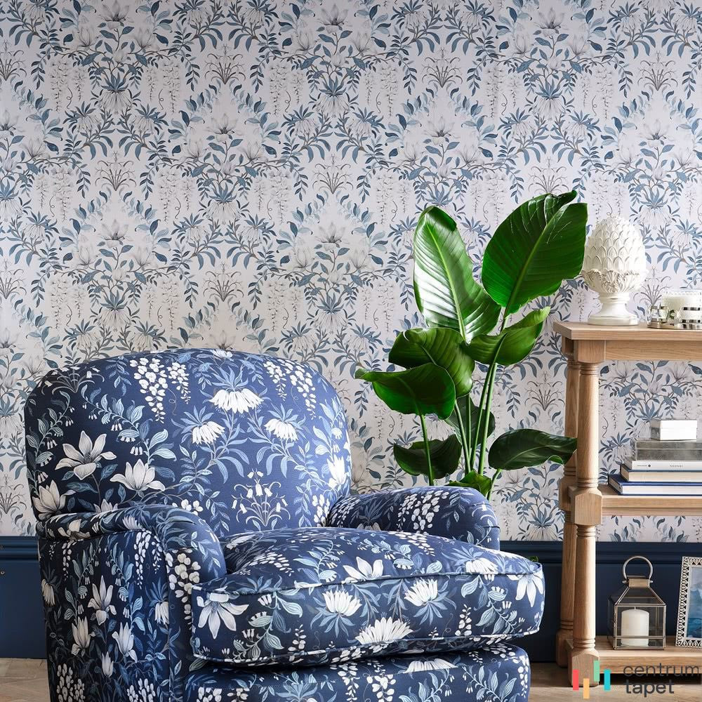 Laura Ashley 113405 za niebieskim kwiatowym fotelem, regałem i zieloną rośliną